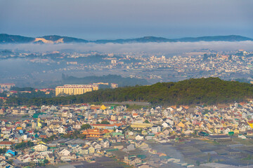 Aerial view of Dalat city with colorful buildings, misty mountains, and lush fields. The serene atmosphere and diverse architecture create a picturesque and tranquil urban landscape.