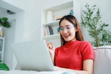 Fototapeta premium Happy young Asian woman using laptop while seated on couch at home