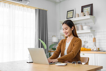 Beautiful young Asian woman working with laptop computer in home kitchen. Work at home