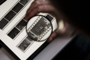 Close-up of a person's hand analyzing film negatives on a lightbox using a magnifying glass to...