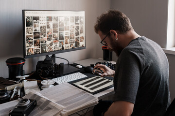 A focused photographer examines film negatives beside a computer displaying a photo collection. A vintage camera and modern equipment surround the workspace.