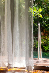 Sunlit sheer curtain in front of glass door, revealing poolside view with lush greenery.