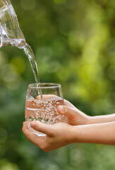 child hands holding glass and water pouring from jug with green garden on the background