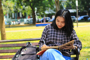 smiling asian beautiful young woman enjoy writing to do list and idea in notebook in outdoors city park