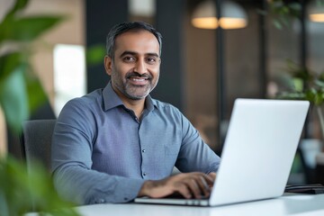 Fototapeta premium Smiling mature business man executive wearing shirt sitting at desk using laptop. Happy busy professional middle aged Indian businessman investor working on computer looking away in office, ai