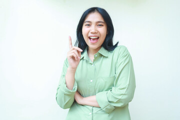 pleased asian young woman wearing green over size clothes pointing finger up with folding arms looking at camera introduce imaginary product isolated on white background