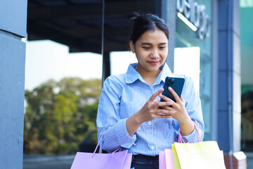 smiling asian woman holding shopping bags and using smartphone feels satisfied while walking in outdoors around mall building