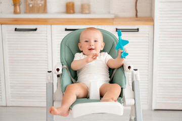 a baby boy eats in the kitchen in a high chair with a spoon, a small child eats on his own