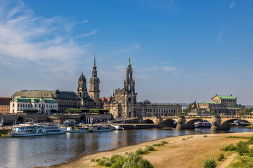 Fototapeta premium Blick auf die Dresdner Altstadt mit der Katholischen Hofkirche (auch bekannt als Kathedrale St. Trinitatis) im Zentrum. Im Vordergrund fließt die Elbe, auf der mehrere Schiffe zu sehen sind. 