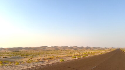Desert road winds through evening dunes and small bushes under the fading light of sunset. Warm evening glow illuminates the path, offering a peaceful journey through the quiet, expansive desert.