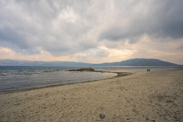 A tranquil scene at Lake Salda, Turkey, featuring a sandy beach curving into the distance, clear waters, and mountains under a cloudy sky.