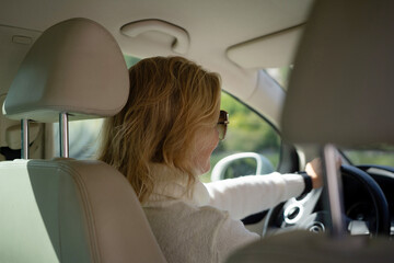 A blonde woman in a white sweater and jeans is driving. Happy woman sitting in a car with a white interior.