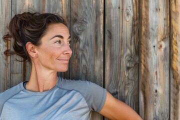 Fototapeta premium A woman leans casually against a wooden wall, exuding a sense of contentment and relaxed elegance in her casual athletic attire, set against a rustic background.