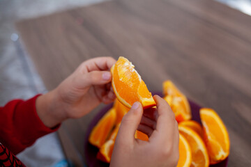 a close-up of a child’s hands holding a slice of orange, with a plate of orange slices on a wooden surface in the background.