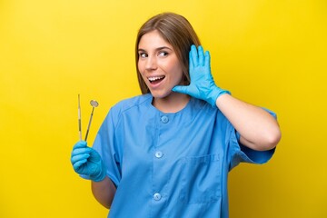 Dentist caucasian woman holding tools isolated on yellow background listening to something by putting hand on the ear