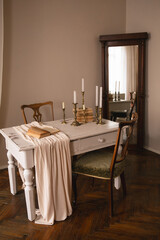 A vintage table draped with beige fabric, brass candlesticks, and an open book. A large wooden mirror reflects the serene, nostalgic room filled with classic furniture and warm tones.