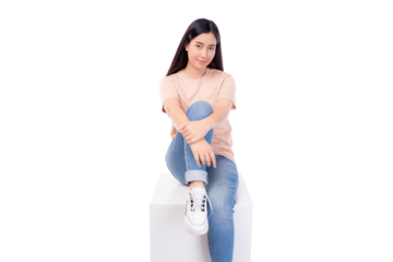 Young Asian woman sitting casually on a white cube, dressed in a light peach t-shirt and jeans, with a friendly and approachable expression looking at camera