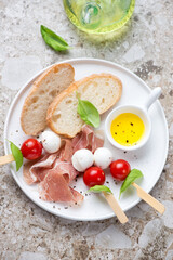 White plate with ciabatta, slices of prosciutto and caprese skewers, vertical shot on a light-brown granite background, high angle view