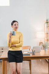 A woman in a yellow sweater holds a coffee cup while standing in a home office setting, taking a work break.