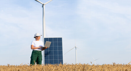 Woman farmer wearing white cap and t-shirt with laptop stands next to solar panel. Wind turbines in...