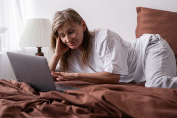 Relaxed middle aged woman lying on bed at home, using laptop, typing on computer keyboard, chatting on dating website. Working in the bedroom.