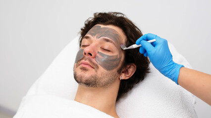 Beautiful caucasian man is doing a cosmetic procedure. Beauty master applies a rejuvenating mask to the male clients face. Skin lifting. The concept of beauty and skincare. Copy place. Close up