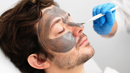Beautiful caucasian man is doing a cosmetic procedure. Beauty master applies a rejuvenating mask to...