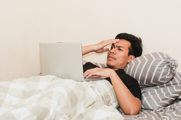 A Southeast Asian man in bedroom and laptop for work from home, serious or thingking in bed for freelance idea. Technology, entrepreneur and remote worker for online job.