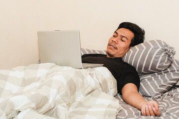 Always online. A Southeast Asian man working on laptop in bed, copy space. Early morning. Pleasant awakenin