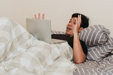 Surprised and shocked A Southeast Asian man is working with his laptop on his cozy bed. Concept of successful freelancer lifestyle.