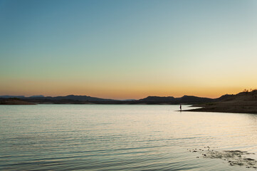 Silhouetted fisherman fishing on lake with desert landscape in background at sunset