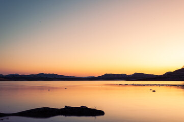 Naklejka premium Tranquil lake with still water reflects the golden light of setting sun against backdrop of distant mountains