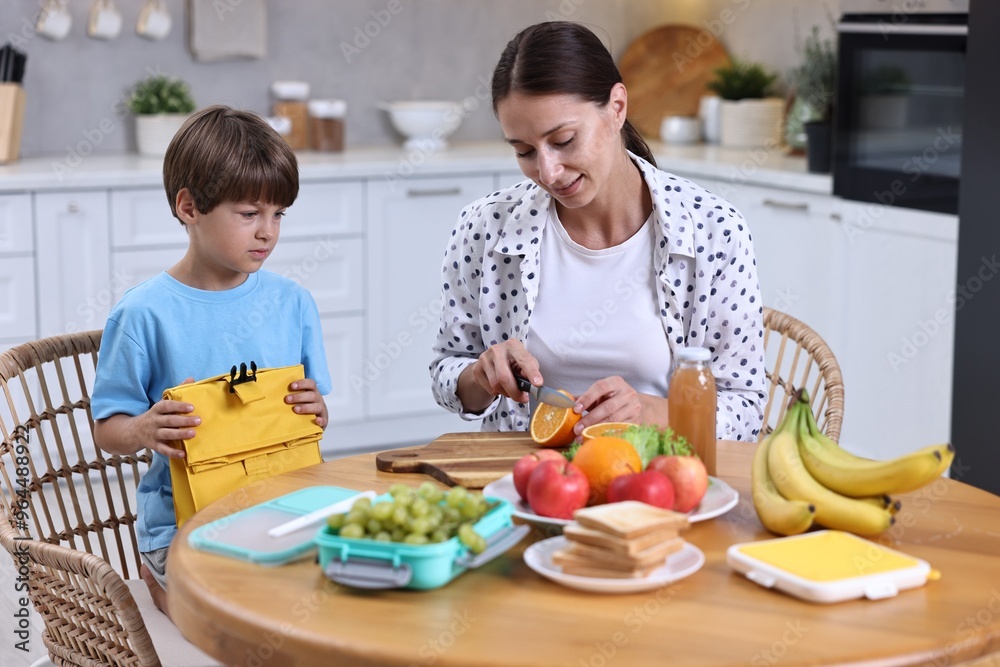 Poster Mother and her cute son preparing lunch box with healthy food at wooden table in kitchen