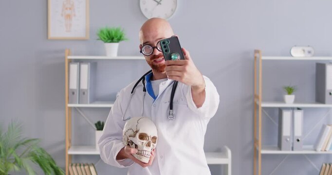 Happy man doctor or nurse captures a funny selfie using a phone, holding a skull in hand. The lighthearted moment unfolds in the hospital office, adding a touch of humor to social media feeds.