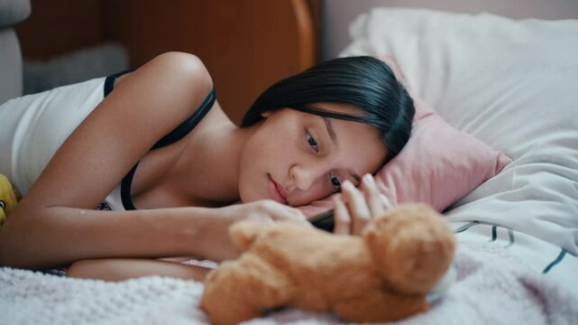 A teenage girl lies lazily on her bed, scrolling through social media on her phone. The room is calm and quiet, with the soft glow of the screen reflecting on her face.