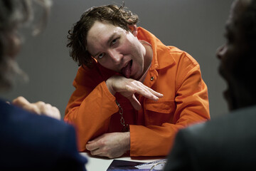 Caucasian man dressed in orange jumpsuit sitting at a table with head slightly tilted, sticking out tongue and pointing finger, expressing defiance and attitude representation