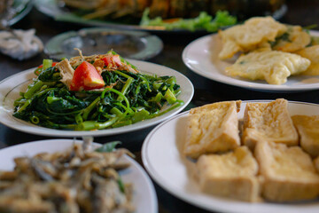 Cah kangkung or Stir fried water spinach is ready to eat on white plate. Surrounded by fried fish and fried tofu. Asian local cuisine and Indonesian food