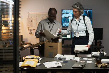 Two professionals analyzing documents while standing in office filled with documents and devices. One person holds documents while other is looking at a file without taking eyes off camera