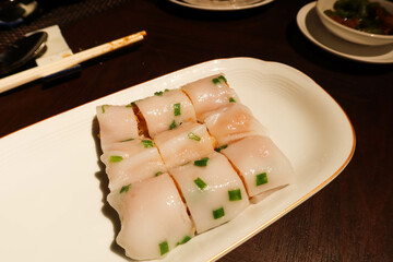 close up of dim sum steamed rice roll with shrimp deep fried crispy spring roll and prawns cut into nine pieces with spring onion as toppings on a white plate pouring soy sauce