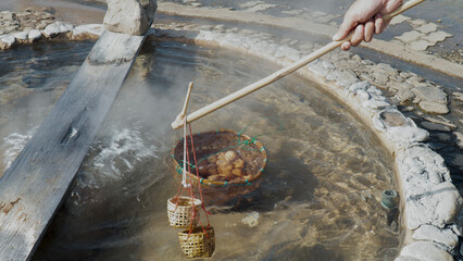 Person boiling eggs in a natural hot spring, using wooden basket and stick. Outdoor thermal cooking process in natural environment.