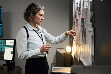 Female detective with short hair analyzing crime scene evidence on bulletin board in office...