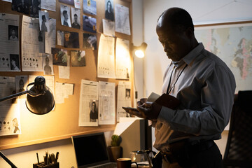 Middle-aged African American man attentively examining notes on bulletin board under soft lamp...