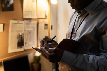 Man taking notes in office while reviewing documents on wall with focused expression and office lit warmly by desk lamp