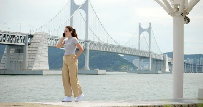 Woman strolls back and forth at Busan's quiet waterfront, engaged in phone call, against beautiful bridge view. Slow-motion, full-length shot captures her relaxed conversation