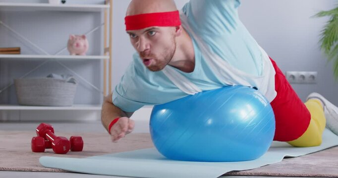 Funny man wearing retro sports clothes doing core strength exercise on fit ball moving arms as if front crawl freestyle swimming with stubborn determined expression on face. Fitness workout concept