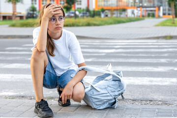 Upset schoolgirl by the road. Sitting at the crosswalk with a backpack. School background.