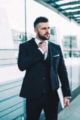 Contemplative businessman in formal wear fixing tie and looking away standing near office building in downtown, serious pensive male banker with laptop computer caring about own appearance