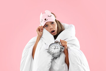 Overslept girl with sleep mask and alarm clock wrapped in blanket on pink background