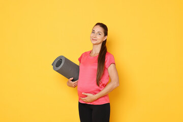 Beautiful pregnant woman with exercise mat on yellow background