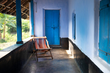 Traditional Kerala home veranda with vibrant blue doors and wooden chair, bathed in sunlight. Rustic charm and lush greenery capture the essence of peaceful village life in rural India.